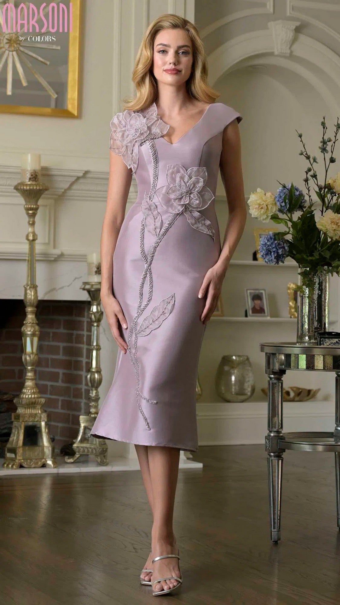 A woman stands indoors near a fireplace and a table with flowers, wearing the Marsoni MV1386 knee-length semi-formal dress in dusty rose.